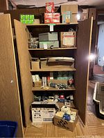 Full view of tall wooden cabinet with shelves filled with various light bulbs in boxes, electrical boxes, and metal lockable boxes as well as small electrical parts in clear containers and boxes.