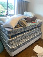 Photo of townhouse stacked twin size mattress with wooden legs, feather pillows, duvet, pillowcases, and folded bedding on a couch near window.