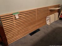 Full view of the Mandal king size headboard laid against a wall showing the horizontal wooden slats and six attached white shelves on one side, set on carpeted floor.