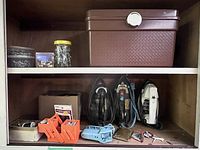 Medium shot of wooden wall cabinet with three vintage irons on middle shelf and orange storage containers with sewing supplies, a brown box on top shelf.