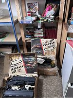 Two open vintage Aurora Thrill Show slot car race track sets with black track sections and white controllers, boxes with colorful car images and instructions, vintage Barbies and puzzles on shelving behind