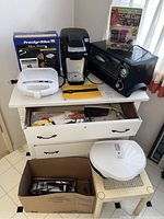 Full lot overview showing all appliances on a white drawer unit and nearby table, includes boxed Proctor-Silex Meal Maker, Keurig coffee maker, toaster oven, slicers, hand mixer, and George Foreman grill.