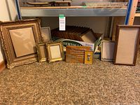 Wide shot of all frames on floor against a metal rack shelf showing nine decorative, wood look, brass and plastic frames in various sizes.