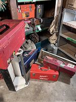 Multiple metal and wooden toolboxes stacked and placed on floor and shelf. Several leather tool belts hanging from a metal rack. Visible labels include Milwaukee on red toolbox.