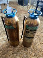 Two vintage brass soda-acid fire extinguishers with blue handles and hoses shown upright on carpeted floor.