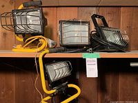 Five assorted work lights placed on shelf against wood panel wall. Lights have metal wire protective guards and yellow or black support frames with power cords.