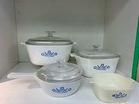 Four Corning Ware Blue Cornflower cookware pieces arranged on white shelf including two casseroles with lids, a saucepan with lid, and a small dish.
