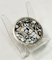 Front-angle photo showing the detailed inlaid mosaic design on the round silver brooch or pendant, displayed on a clear stand.