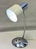 Front side view of chrome and tin gooseneck desk lamp, lit with spiral CFL bulb inside metal shade.