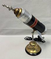 Side angled view of vintage rocket table lamp showing black body with metallic silver tip, fins, and brass base with rivets.