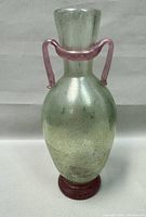 Front view of a greenish speckled blown glass vase with two curving pink glass handles and a pink base.