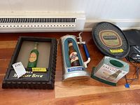 Four plastic beer and bar lights on wooden floor near baseboard heater, various shapes and sizes including Miller Brew black frame, Sterling beer stein, Champale rectangular light, National round tray