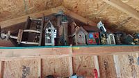 Wide angle photo showing an assortment of wooden and plastic birdhouses and feeders placed on a high shelf inside a shed.