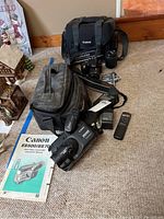 Photo showing Nikon D3300 camera with Olympus strap, Canon black camera bag, Canon ES500 camcorder, tripod, remote control, and instruction manual spread on carpet.