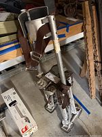 Pair of aluminum drywall stilts, showing top leg cuffs with brown adjustable straps and metal buckles, the padded leg supports and foot platforms with spring mechanism visible.