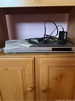 Silver Toshiba DVD player on wooden cabinet with remote control, power adapter and manual on top.