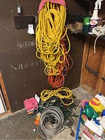 Photo of yellow heavy-duty extension cords hanging from a wall, orange cords mixed in, and coiled green and gray extension cords on the floor in a garage corner with various cleaning supplies and electrical items nearby.