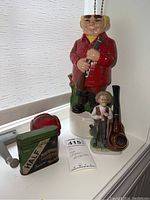 Photo showing large ceramic pipe holder figurine, small ceramic pipe holder figurine, vintage tobacco tin, and wooden pipe on white surface.