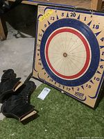 Photo showing the dart board and pair of men's ice skates placed on floor with green carpet and concrete basement floor visible.