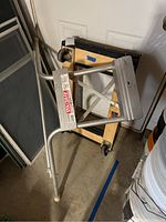 Full view of Sta Safe metal ladder stabilizer leaning against wall near wood-framed dolly.