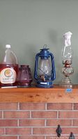 Wide shot showing the oil lamps and lantern along with the gallon jug of lamp oil on a wooden shelf against a light green wall.