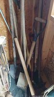 Photo showing several wooden handled garden tools including shovels and a spade leaning against a wall in a shed.