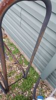 Close-up view of the curved metal bars of the log holder showing rust and weathering against a background of siding and ground.