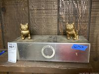 Front view of two gold-colored metal Mack bulldog hood ornaments mounted on a hollow metal rectangular case with a label.