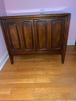 Front view of the closed walnut nightstand showing two recessed-panel doors and tapered legs against a pink wall
