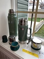 Two green Stanley vacuum bottles with metal lids and plastic seals, placed on a window sill. The larger bottle has a side handle. Both show wear from use.