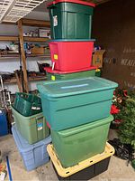 Stack of various plastic storage bins and one green plastic basket in a garage setting.