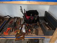 Shelf with laid out tools including crow bar, hand saw, wrenches, screwdrivers, hammers, and cloth tool case.