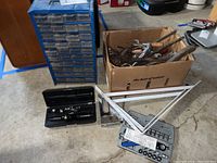 Full lot showing blue hardware bin with many drawers, cardboard box with rusty tools, screwdriver sets in cases, stapler, and measuring squares on concrete floor.