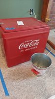 Front view of red Coca-Cola cooler next to galvanized Coca-Cola pail