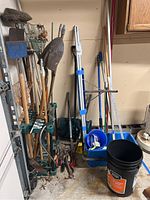 Overview of long-handled garden tools stored in green plastic tool rack including shovels, rakes, brooms, a dustpan, bucket, and other tools beside the rack. Tools have dirt and wear, set in a garage environment with concrete floor and some debris.