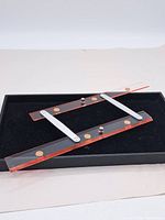 Top view of the clear acrylic adjustable ruler with red edges, metal pivot arms, and brass screws resting on a black surface.