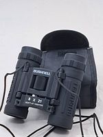 Full view of black compact Bushnell binoculars with strap and soft carrying case behind.