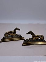 Pair of brass bookends with reclining greyhound dog figures on stepped bases, viewed from front and side.