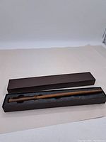 Full view of wooden wand in black foam inside rectangular brown box with lid.