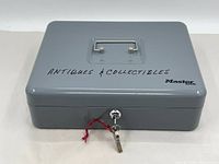 Front view of gray metal locking money box by Master Lock with 'Antiques & Collectibles' handwritten on lid and keys in lock.
