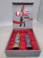 Two limited edition Diet Coke bottles in an open display case with red interior lining and Diet Coke branding on lid.