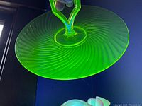 Close-up photo under UV light showing bright green glow of uranium glass plate and central handle, highlighting the spiral pattern of the plate.