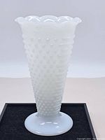 Side view of opaque white hobnail milk glass vase with ruffled rim on black surface.