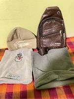 Side view of tan knitted hat, two fleece scarves and small brown shoulder bag on red and orange checked surface
