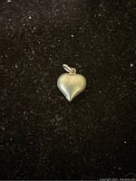 Photo of a small silver heart-shaped locket pendant against a black background. The locket has a loop for a chain and a smooth matte finish.