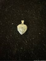 Close-up of a silver heart-shaped locket pendant on a dark background showing leaf pattern engraving.