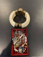 Photo showing a handmade ivory resin cuff with metal bands beside a small box filled with assorted costume jewelry pieces such as necklaces, brooches, and earrings.