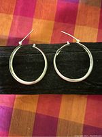 Pair of silver hoop earrings photographed against a black velvet background on a colorful fabric surface, showing their shape and size.
