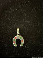 Close-up image of small silver horseshoe pendant with red and green enamel dots on a dark textured cloth background.