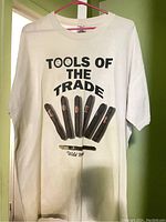White XL cotton T-shirt with 'TOOLS OF THE TRADE' text and graphic of cigars and a cigar cutter on front, hanging on pink hanger against beige wall.